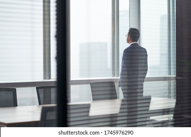Rear Side View Of An Asian Business Man Standing In Front Of Office Window Looking Out And Thinking Hands In Pockets