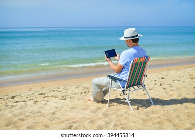 Rear Side View Om Man Using Digital Tablet Relaxing On Chair At The Ocean Beach. Blue Sky Outdoors Background