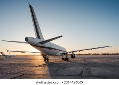 Rear three quarter view of passenger airplane on airport apron and few other planes in the background. Low evening sun creates a long shadow of aircraft silhouette on concrete surface. 