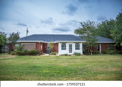 The Rear View Of A 1980's Style Red Brick Ranch House With White Siding And A Large Backyard