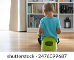 Rear view of a 3-year-old Caucasian boy in light blue t-shirt sitting on green potty reading book, large bookcase full of books in background. Potty training concept. Love reading concept. Copy space