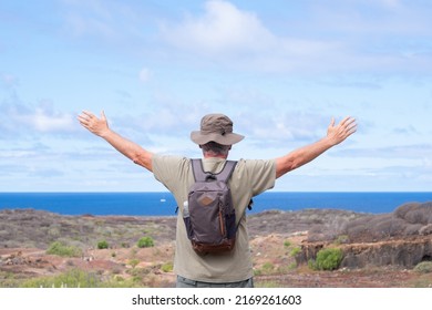 Rear View Of Active Senior Man With Backpack And Hat While Hiking Outdoors Looking At The Horizon Over Sea With Outstretched Arms. Elderly Man Enjoying Retirement And Healthy Lifestyle.