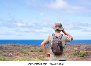 Rear View Of Active Senior Man With Backpack And Hat While Hiking Outdoors Looking At The Horizon. Elderly Man Enjoying Retirement And Healthy Lifestyle. Blue Sea And Sky In The Background