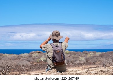 Rear View Of Active Senior Man With Hat And Backpack In Outdoor Excursion Looking At The Horizon Over Sea And Pointing At The Big Cloud In Front To Him