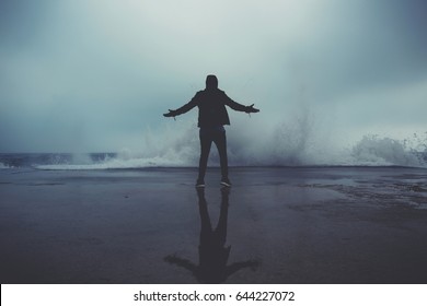 Rear view of a adult man standing with arms raised in front of the sea despite a great wave hits the shore, young male enjoying breathtaking scenery of the sea at cool overcast rainy day