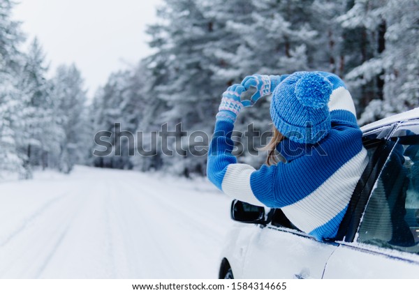 Rear view of adult woman in car over\
snowy forest on winter roadtrip. Wanderlust concept. Winter\
travelling scene. Making shape of love heart by\
hands.