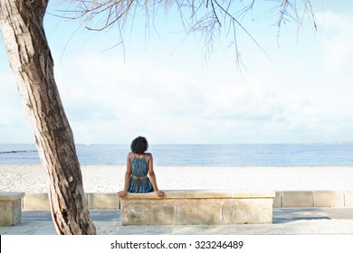 Rear view of african american black young woman sitting on the edge of a white sand beach contemplating the sea on a sunny holiday destination, outdoors. Travel lifestyle and healthy living, exterior. - Powered by Shutterstock