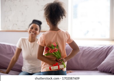 Rear View African American Daughter Holding Flowers, Bouquet And Gift Behind Back, Congratulate Young Diverse Mother With Mothers Day Or Birthday, Adorable Child Making Surprise To Mum At Home.