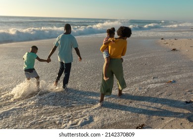 Rear View Of African American Family Wading On Shore At Beach During Sunset. Unaltered, Family, Lifestyle, Togetherness, Enjoyment And Holiday Concept.