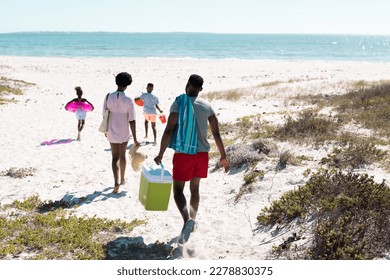 Rear view of african american family with swimming float, cooler walking at beach towards seascape. Copy space, unaltered, parents, together, childhood, picnic, nature, vacation, enjoyment, summer.