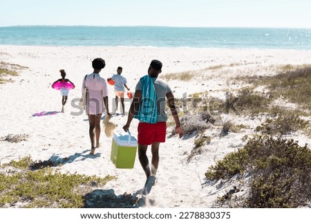 Rear view of african american family with swimming float, cooler walking at beach towards seascape. Copy space, unaltered, parents, together, childhood, picnic, nature, vacation, enjoyment, summer.