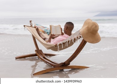 Rear View Of African American Man Reading A Book While Relaxing On A Hammock At Beach