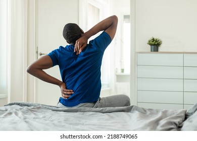 Rear View Of African American Man Touching And Massaging Aching Neck And Spine Suffering From Pain After Sleeping On Uncomfortable Bed Sitting In Modern Bedroom At Home. Arthritis Symptom - Powered by Shutterstock