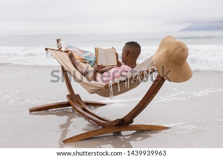 Rear view of African american man reading a book while relaxing on a hammock at beach