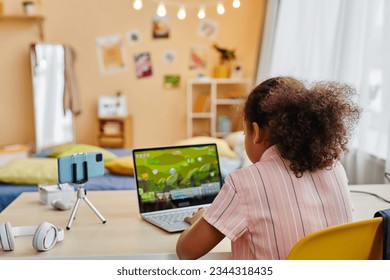 Rear view of African American schoolgirl sitting by desk in front of laptop and smartphone camers and playing video game in home environment - Powered by Shutterstock