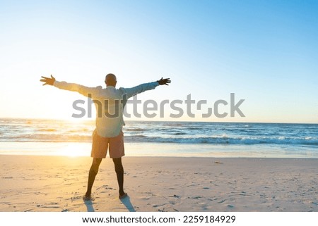 Rear view of african american senior man with arms outstretched looking at seascape against blue sky. Dusk, copy space, carefree, happy, retirement, unaltered, beach, vacation, enjoyment, nature.