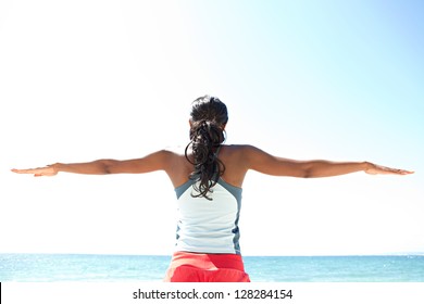 Rear view of an "african american" woman stretching her arms to the sides while standing against a deep blue sky, exercising on a sunny day by the sea.