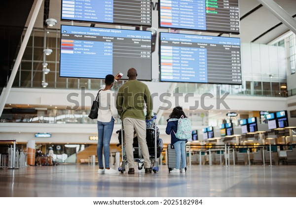 Rear view of\
african family with luggage looking at scheduled flights time table\
at airport terminal. Parents with daughter waiting at airport and\
checking flight scheduled.