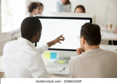 Rear view of african team leader explaining caucasian colleague corporate software usage, black manager helping coworker with computer task, mentor teaching intern, giving instructions about emails