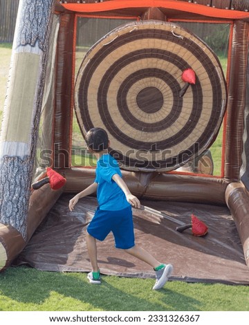Rear view Asian boy throwing realistic plastic axe into inflatable axe game outdoor shooting darts board at park Flower Mound, Texas, USA. Safe realistic Viking throw with vinyl floor tether straps