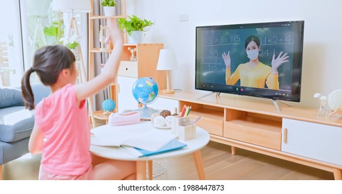 Rear View Of Asian Girl Is Learning Math And Looking Online Course On TV In Living Room - Elementary School Female Teacher Wearing Face Mask While Local Authority Has Announced A Stay At Home Order