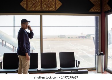 Rear View Of A Asian Man Making A Phone Call While Standing In Front Of A Glass In An Airport Waiting Room