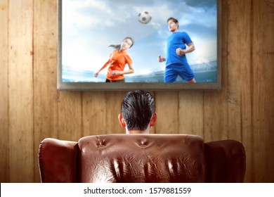 Rear View Of Asian Man Sitting On The Couch Watching A Football Game On Television At Home