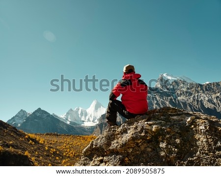 rear view of asian man sitting on top of rock with Yangmaiyong (or Jampayang in Tibetan) mountain peak in the distance in Yading, Daocheng County, Sichuan Province, China