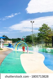 Rear View Asian Toddler Boy Playing At Splash Park Near Dallas, Texas, America. Colorful Recreation Site With Splashing Water Fountains For Kids Summertime Activities