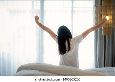 Rear View Of Asian Woman Stretching Hands And Body In Bed After Wake Up In Bedroom At Home. Concept For Start New Day With Happiness. Copyspace On The Right. Young Happy Student Or College Female Life