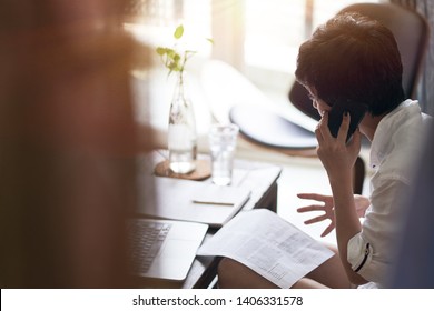 Rear View Of Asian Woman Talking On Smart Phone And Working On Laptop At Home 