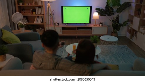 Rear View Of Asian Young Couple Eating Pop Corn Are Watching Green Chroma Key Screen TV And Having Fun While Sitting On Couch At Night In The Living Room