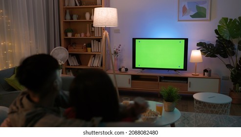Rear View Of Asian Young Excited Couple Are Watching Sport Games On TV With Green Chroma Key Screen And Having Fun While Sitting On Couch At Night In The Living Room