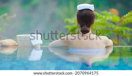rear view of asian young woman lying on the bathtub is relaxing in hot spring with towel on head