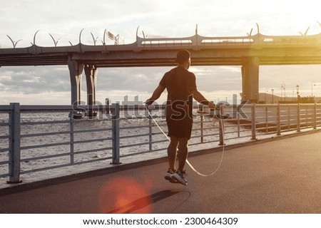Rear view athlete man working out on skipping rope in urban embankment outdoors at sunset background. Sportsman workout with jumping rope in city district. Healthy lifestyle concept. Copy text space