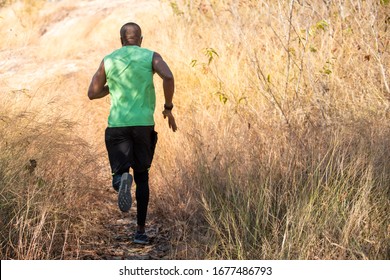 Rear View Of Athlete Trail Runner Sportsman Running Away Uphill On Mountain Trail. African Man In Sport Shoes And Sportswear Sprinting And Endurance Training Outdoors Trail Running Workout.