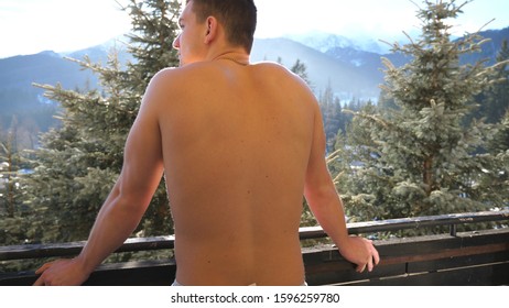 Rear View Of Athletic Guy Stands On Balcony And Breathes Fresh Air During Sunny Day. Young Man Enjoys Beautiful Mountain And Forest Landscape In Morning. Concept Of Winter Vacation Or Holiday. Slow Mo