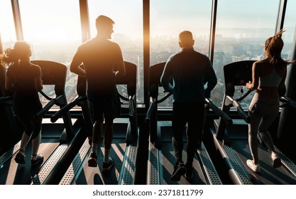 Rear view of athletic people jogging on treadmills in a health club near window with city view at sunset. Silhouettes of young people exercising in a gym with a stunning view.
