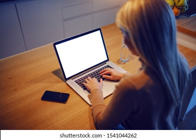 Rear view of attractive blonde woman typing on laptop while sitting at dining table at home. On table are smart phone and glass of water. - Powered by Shutterstock