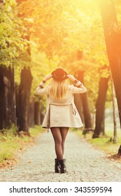 Rear View Of Attractive Young Blonde Woman In Autumn In Park Wearing Beige Trench Coat, Black Boots And Black Fedora Hat. Vibrant Colors, Retouched, Filter Applied.