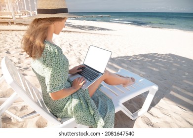 Rear view of an attractive young Caucasian woman in a green dress and hat sits on a lounger with a laptop in her hands in the shade of a wooden umbrella. Cut out laptop screen - Powered by Shutterstock