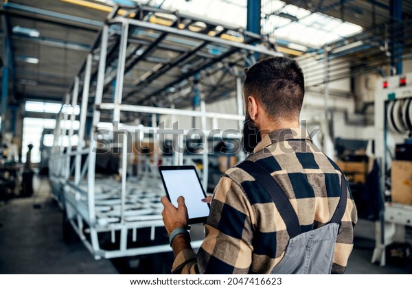 Rear view of auto industry\
worker standing in a factory with tablet in his hands and looking\
at metal construction prepared for a new bus. Auto-mechanic in bus\
factory