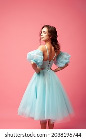 Rear View Of The Back Of A Fragile Young Woman In A Blue Evening Dress With A Full Skirt And Lantern Sleeves, Isolated On A Pink Background. 