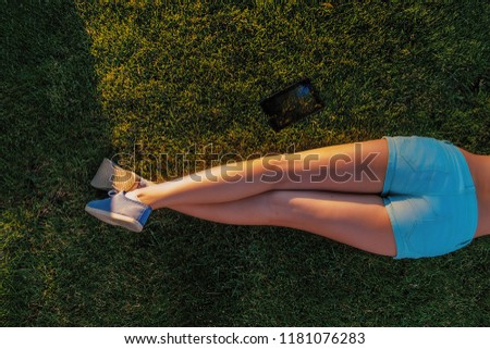 Rear view of bare legs of lying girl on grass in teal shorts and canvas trainers and tablet computer lying near on grass, backlit by sun