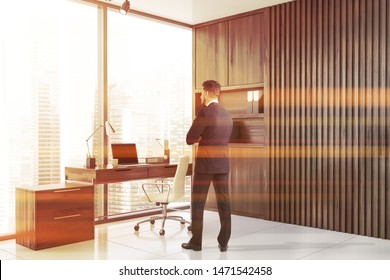 Rear view of bearded businessman standing in modern CEO office with wooden walls, tiled floor, wooden computer desk and bookcase with folders. Concept of leadership. Toned image