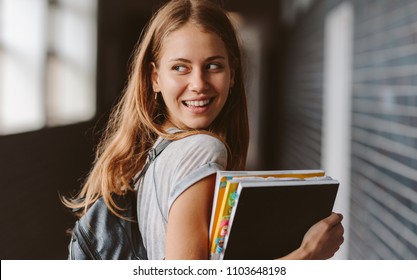 Rear View Of Beautiful Female Student Walking Through College Hallway Looking Back And Smiling. Girl Going For The Class.