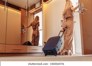 Rear view of a beautiful slim young woman in brown stylish overalls with suitcase leaves the hotel room after long therapeutic vacation. Young business woman goes on a business trip leaving her house