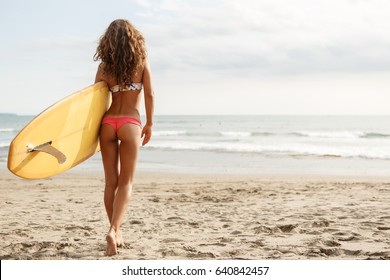 Rear view of beautiful surfing girl in sexy red bikini with sporty butt hold yellow surf longboard surfboard board on a beach at sunset or sunrise. Modern active sport lifestyle and summer vacation.