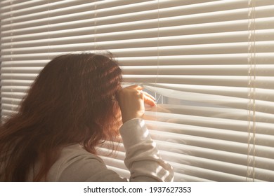 Rear View Of Beautiful Young Woman With Long Hair Peeks Through Hole In The Window Blinds And Looks Out The Window. Surveillance And Curiosity Concept