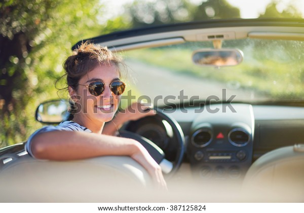 Rear view. Beautiful young woman happy to drive her\
convertible car on a country road in summer. she looks at camera.\
Shot with flare